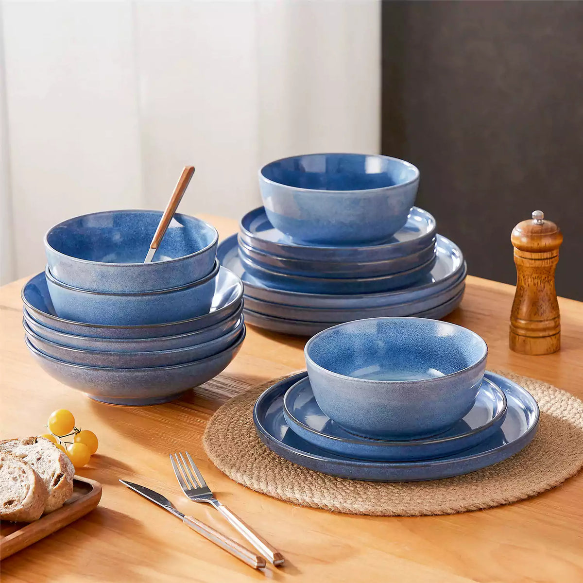 Anya 16-piece blue stoneware dinnerware stacked on a light brown wooden table with bread and woven placemats.