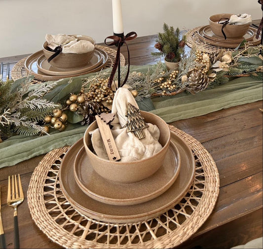 Anya 16-piece caramel dinnerware set on a Christmas table with woven bamboo placemats, candle holders, green table runner, and festive decorations.