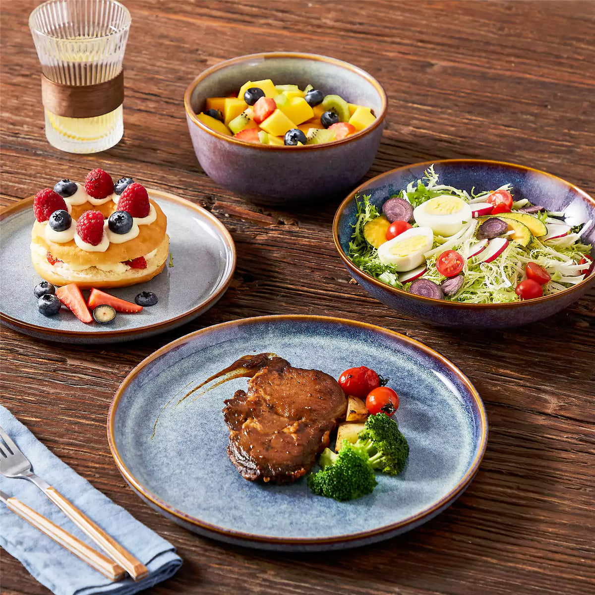 laya 16-piece deep indigo stoneware dinnerware set on a dark walnut wood table, served with roasted lamb chops, soufflé, fruit bowl, and green salad.