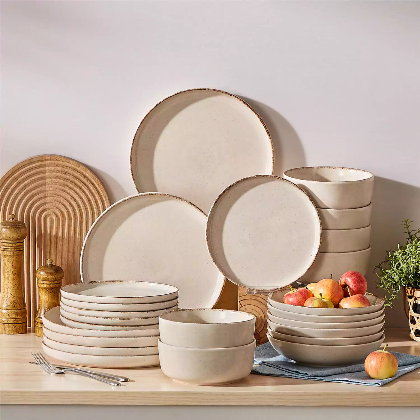 Plays 24-piece stoneware dinnerware set neatly stacked on a kitchen stovetop, styled with green plants, fresh apples, and a blue napkin for a modern everyday table setting.