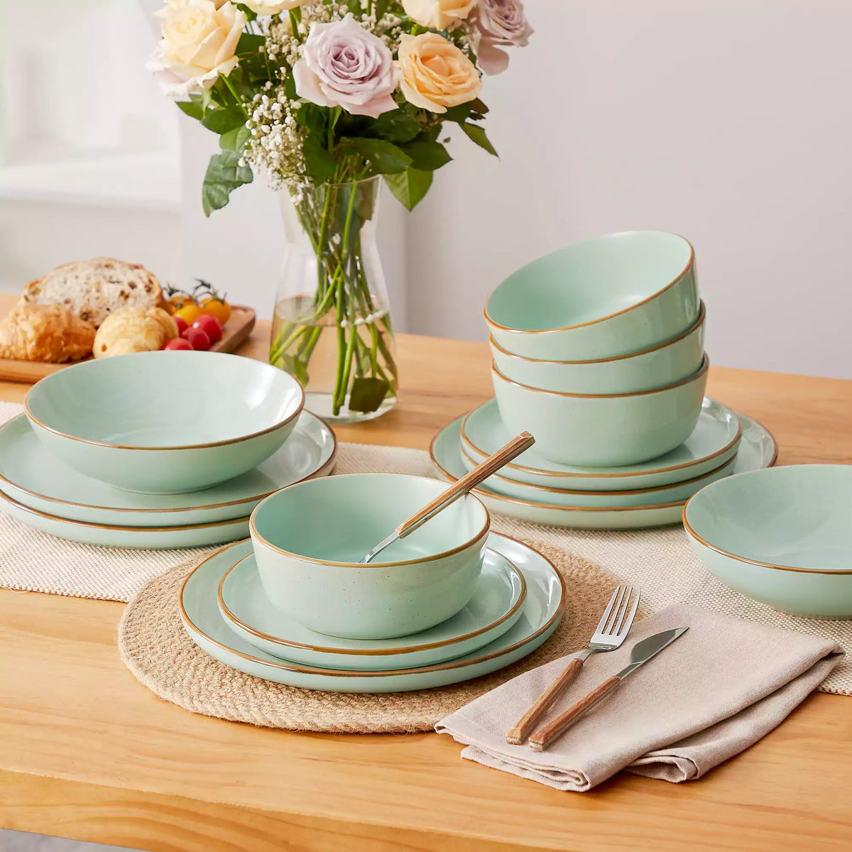 Green rustic stoneware dinnerware set with speckled lipped plates and bowls on a wooden dining table with flowers, napkins, and bread.