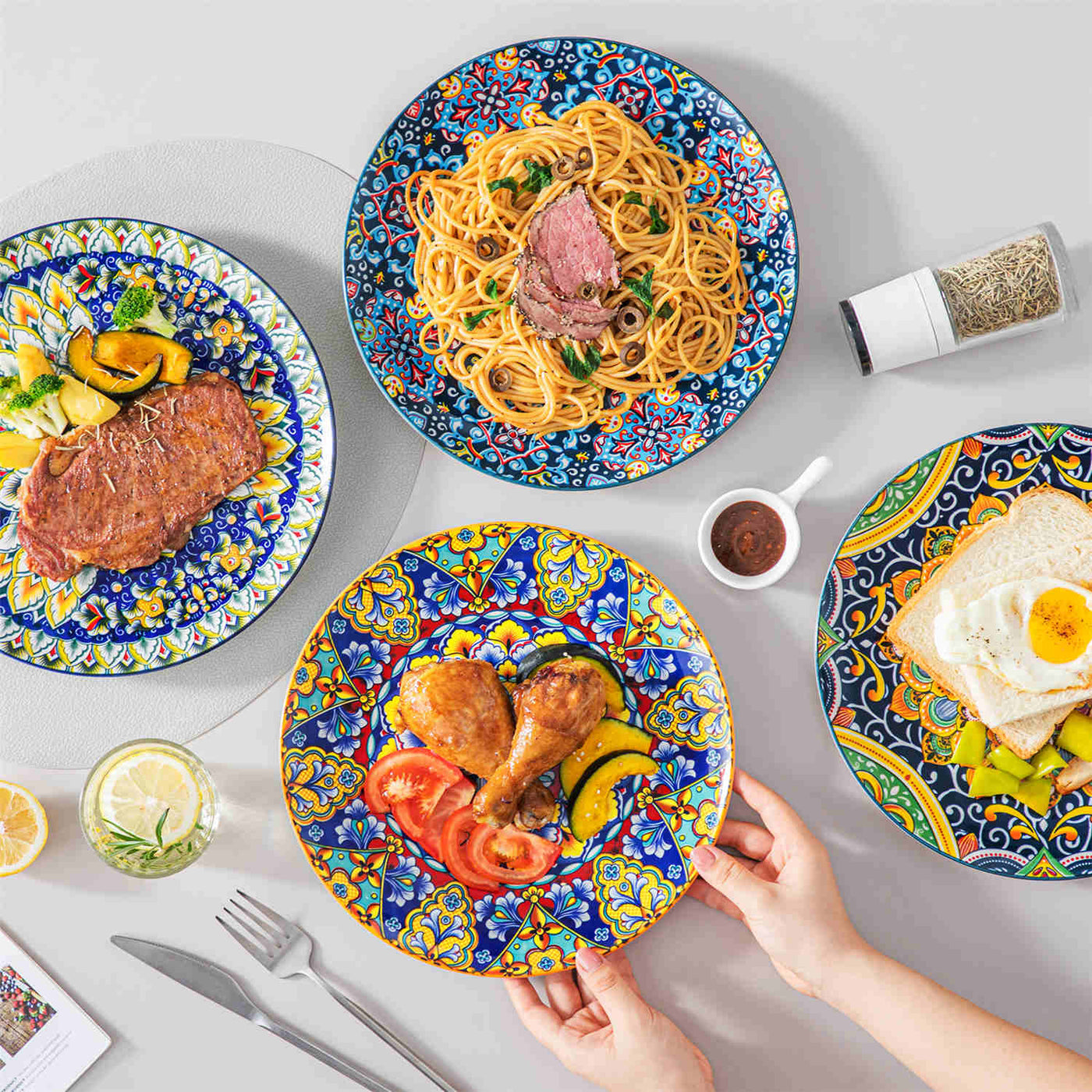 Boho-style porcelain dinner plate set with vibrant floral patterns, serving steak, pasta, chicken, and toast on a grey-white table setting.