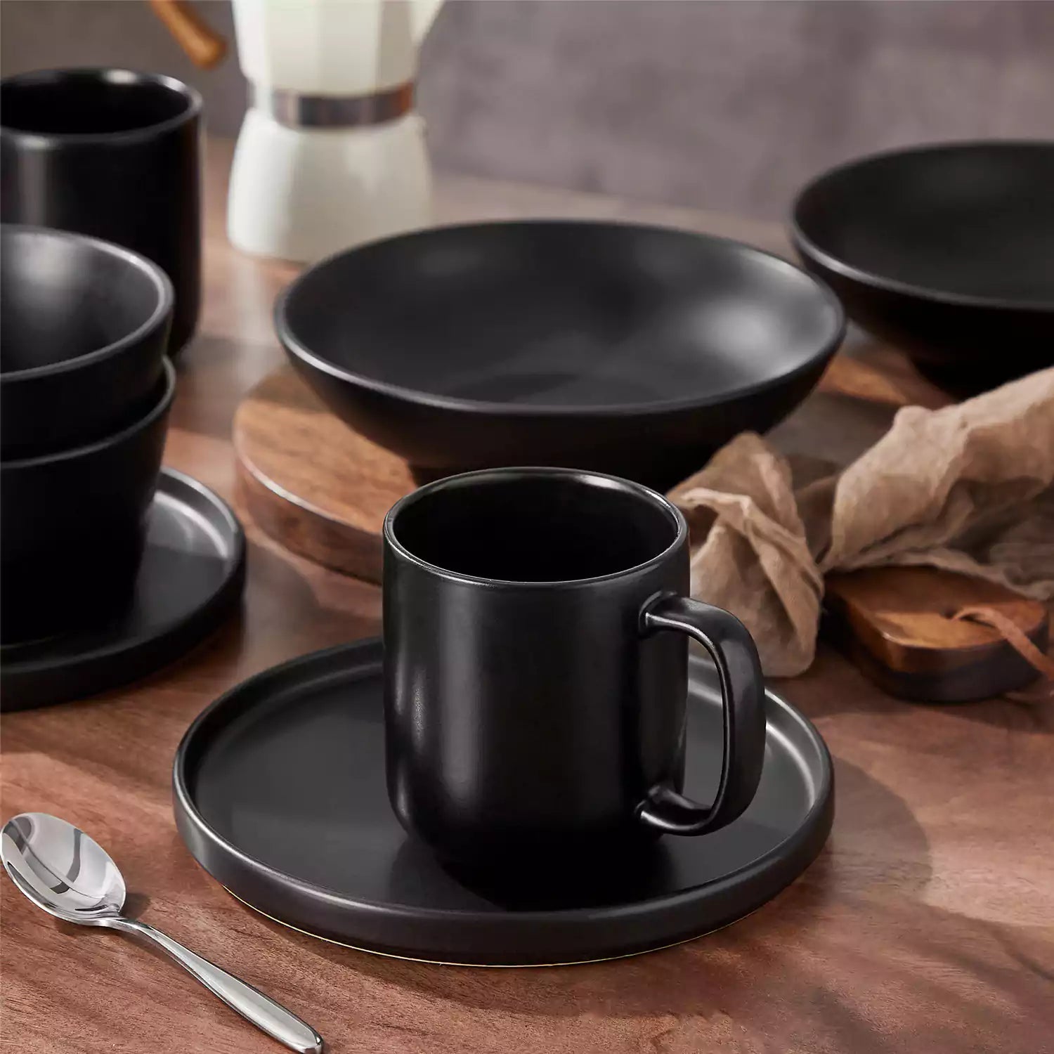 Playa 16oz stoneware mugs in black reactive glaze on a rustic wooden café table with coffee maker and pastries