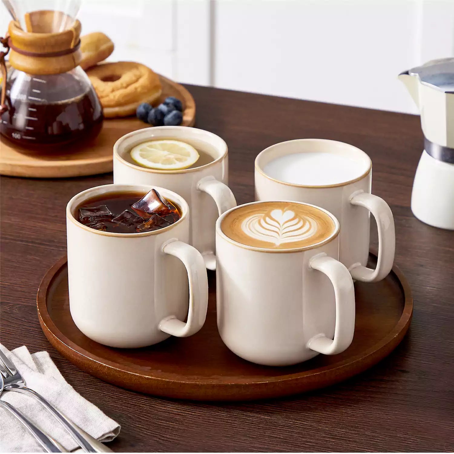 Playa 16oz stoneware mugs in beige reactive glaze on a rustic wooden café table with coffee maker and pastries