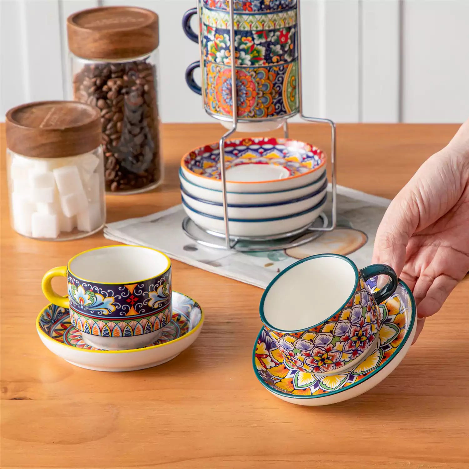 Simi 12 piece porcelain cafe set with 5oz flat white cups and saucers in vibrant boho chic floral patterns on a black metal storage rack with a magazine and napkin on a wooden table