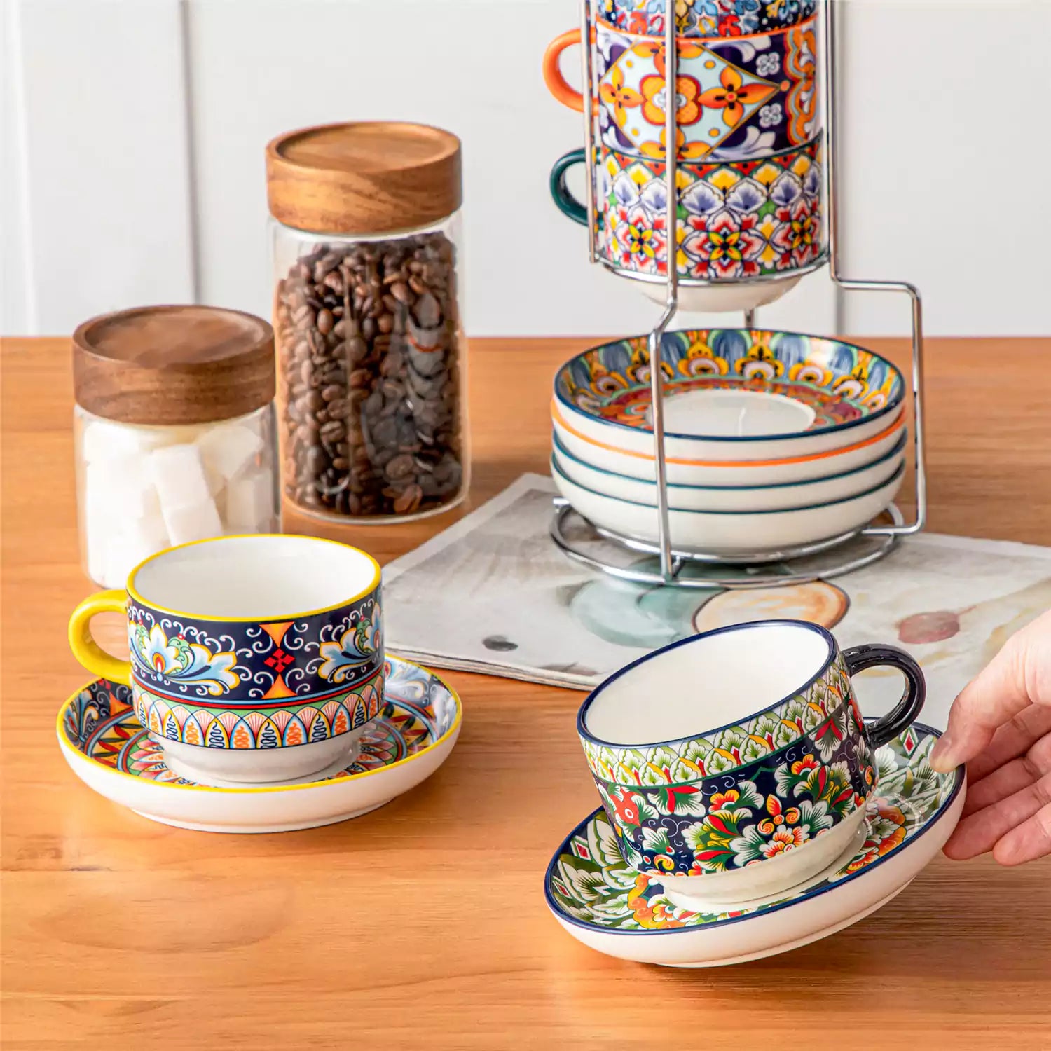 Simi 12 piece porcelain cafe set with 9oz flat white cups and saucers in vibrant boho chic floral patterns on a black metal storage rack with a magazine and napkin on a wooden table