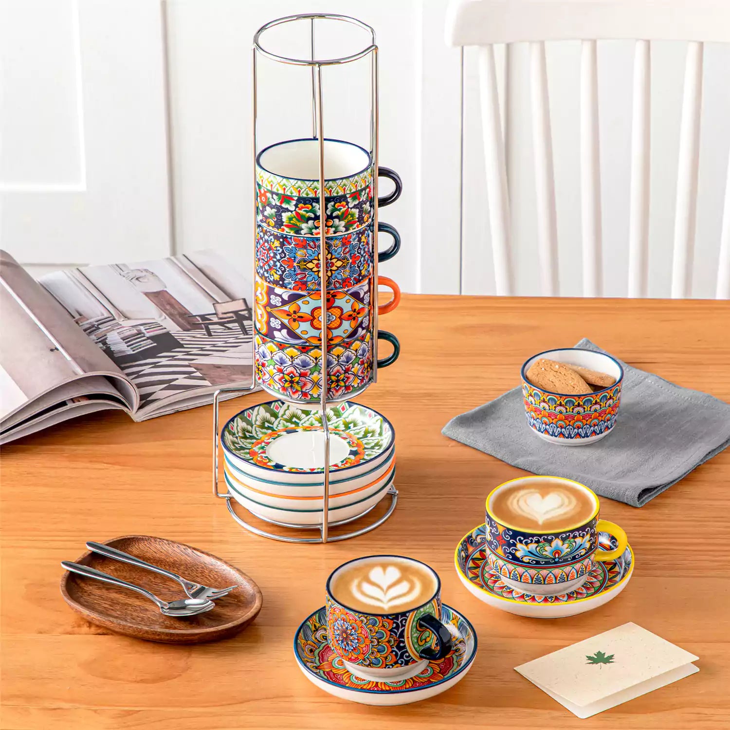 Simi 12 piece porcelain cafe set with 9oz flat white cups and saucers in vibrant boho chic floral patterns on a black metal storage rack with a magazine and napkin on a wooden table