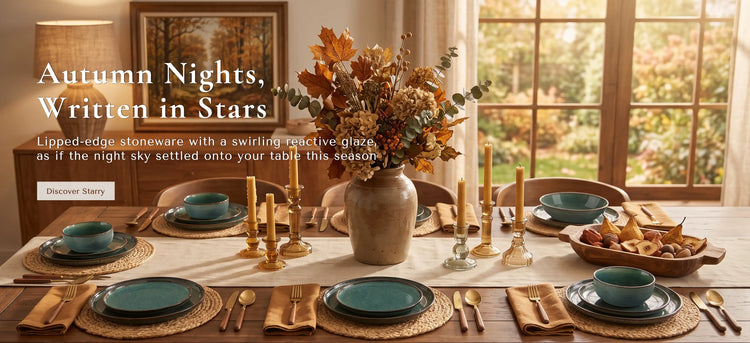 An autumn-themed dining table setting in a warm, sunlit room. The table is styled with teal ceramic bowls, gold-toned cutlery, woven placemats, and a large central vase filled with fall foliage.